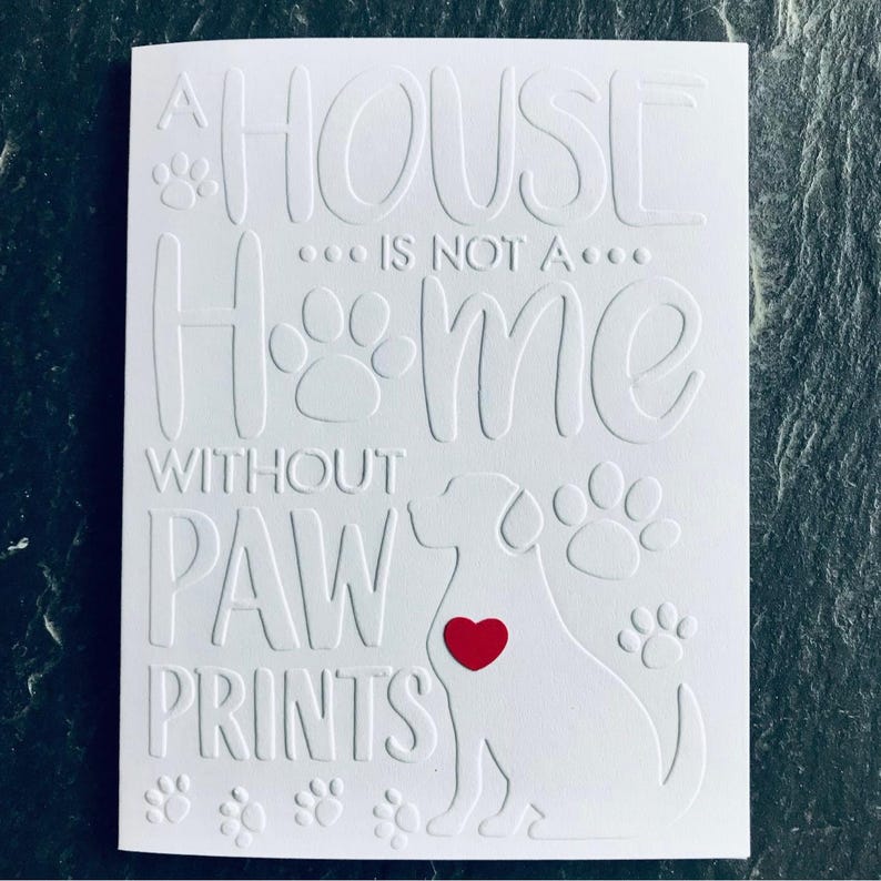 Puede incluir: Tarjeta blanca con el texto "Una casa no es un hogar sin huellas de patas" y una silueta de perro con un coraz&oacute;n rojo en el centro. El texto est&aacute; en relieve y la silueta del perro est&aacute; recortada.