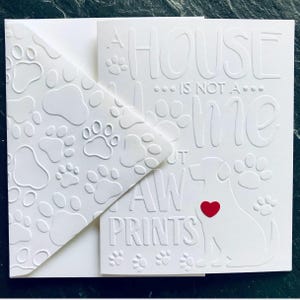Puede incluir: Una tarjeta blanca con un patr&oacute;n de huellas de patas en el sobre. La tarjeta dice "Una casa no es un hogar sin huellas de patas" con un coraz&oacute;n rojo y una silueta de perro.