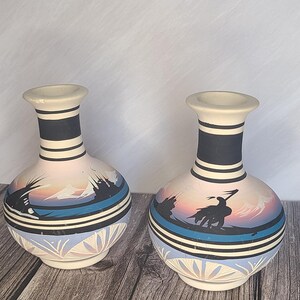 May include: Two matching ceramic vases with a matte cream finish, featuring a hand-painted landscape scene in black, blue, and soft pink. The design includes horizontal black bands on the neck and base, with a silhouette of a figure on horseback.