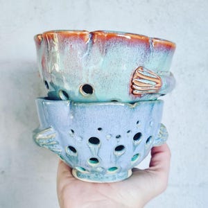 May include: Two handmade ceramic bowls stacked. The top bowl is aqua with an orange rim, the bottom is light blue. Both have decorative holes and fish-shaped handles. The bowls are likely for decorative use.