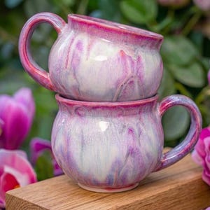 May include: Two ceramic mugs stacked on top of each other. The mugs are a rounded shape with a handle and are glazed in white, purple, and pink. The mugs have a glossy finish. They are on a wooden surface, with pink flowers in the background.