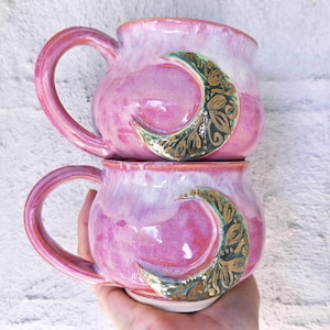 May include: Two stacked ceramic mugs featuring a glossy pink and white marbled glaze. Each mug displays a prominent crescent moon shape on the side, decorated with intricate gold-toned floral patterns and filigree details against a dark base.