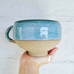 May include: A large, handmade ceramic mug with a light blue glaze and a beige rim. The mug has a rounded shape and a single handle.