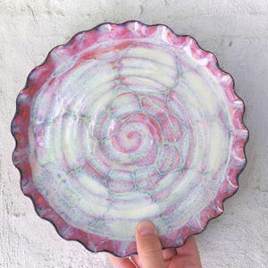 May include: A handmade ceramic plate with a frilled edge. The plate features a spiral design in shades of pink, white, and blue. The rim is a darker pink with a black edge. The plate is held by a hand.