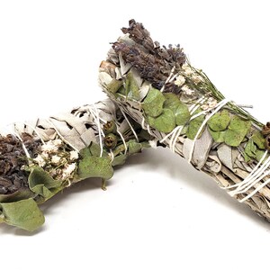 May include: Two sage bundles, each wrapped with white string. One bundle features dried lavender and white flowers, while the other has green eucalyptus leaves and small brown seed pods. The bundles are on a white background.
