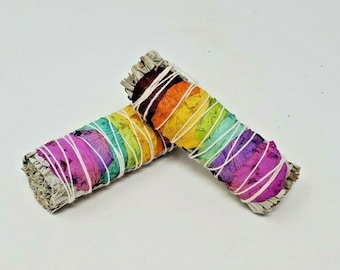 May include: Two rainbow smudge sticks with white string wrapped around them. The sticks are made of dried herbs and flowers.