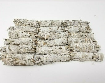 May include: A stack of white sage smudge sticks, tied with white string, on a white background.