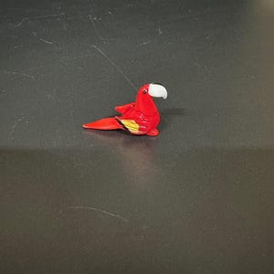 Puede incluir: Una pequeña figura de vidrio de un loro rojo con un pico blanco. El loro está posado en una rama con las alas extendidas.