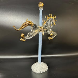 Puede incluir: Una figurita de unicornio de vidrio con detalles dorados, montada en un poste azul y blanco con una base de vidrio transparente.