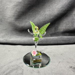 May include: A glass fairy figurine with green wings and a delicate, transparent body. The fairy stands atop a mirrored base with a small, square, mirrored box adorned with pink and white flowers. The fairy's wings are a vibrant green.