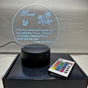 Puede incluir: Lámpara LED redonda de acrílico transparente con la palabra "Mom" y un mensaje de amor grabado. La lámpara presenta un diseño de mariposa y rosas. Se incluye un mando a distancia para ajustar el color. La lámpara se apoya en una base negra.