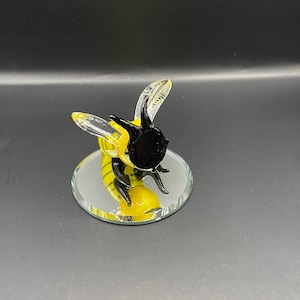 Puede incluir: Una figurita de vidrio de una abeja con un cuerpo rayado amarillo y negro y alas transparentes. La abeja está de pie sobre una pequeña base redonda de espejo.