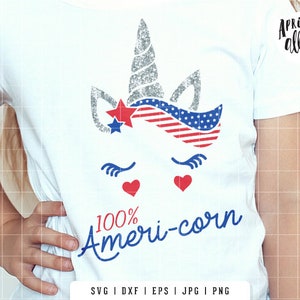 May include: White t-shirt with a unicorn graphic in patriotic colours. The unicorn has a silver glitter horn and ears, with a mane and star accents in red, white, and blue. The text "100% Ameri-corn" is written in blue script.
