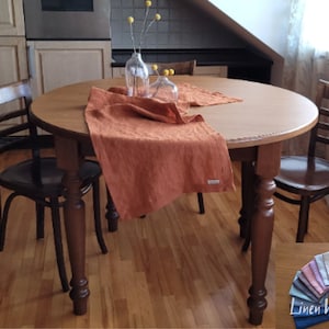 Puede incluir: Una mesa redonda de madera con dos sillas y un camino de mesa de lino de color naranja quemado. La mesa tiene patas de madera torneadas. El camino de mesa es de lino y tiene una textura sutil. La mesa está puesta con un jarrón de flores secas.