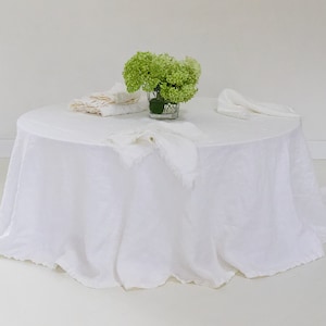 May include: A white linen tablecloth with a ruffled edge, set on a round table. A small vase with green flowers sits in the center of the table, surrounded by folded napkins.