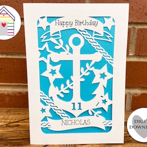 May include: A white card with a blue background and a white anchor design. The anchor is surrounded by stars and a banner that says "Happy Birthday". The number "11" and the name "Nicholas" are also on the card.