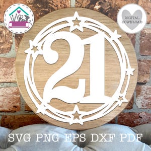 Puede incluir: Un letrero redondo de madera con el número 21 recortado en blanco, rodeado por un diseño de estrellas y remolinos blancos. El letrero tiene un fondo de vetas de madera marrón claro. Las palabras "SVG PNG EPS DXF PDF" están en la parte inferior.