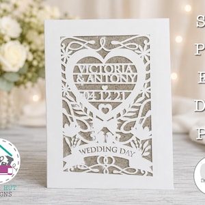 May include: A white laser-cut card featuring a central heart design with the text VICTORIA & ANTONY and 04.12.21, surrounded by floral and vine patterns. The bottom banner displays the text WEDDING DAY above two interlocking rings.