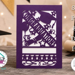 May include: A purple laser-cut greeting card featuring the word CONGRATULATIONS, a graduation cap, a diploma, stars, and a balloon. The design includes a horizontal band of stars and decorative swirls at the bottom, set against a white background.