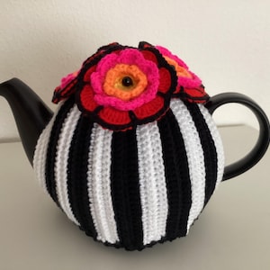 Op de afbeelding: Een zwart-wit gestreepte theepot met een gehaakte hoes. De hoes is versierd met felroze, oranje en rode gehaakte bloemen. De theepot heeft een zwart handvat en tuit.