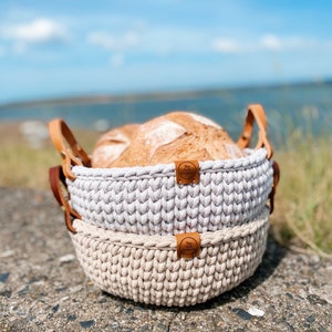 May include: Two crocheted baskets with leather handles, one gray and one beige, holding a loaf of bread. The baskets have a brown leather tag with the text "The Loft Bakery" on them.