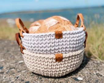 Könnte beinhalten: Zwei gehäkelte Körbe mit Ledergriffen, einer grau und einer beige, die ein Brot halten. Die Körbe haben ein braunes Lederetikett mit dem Text "The Loft Bakery" darauf.