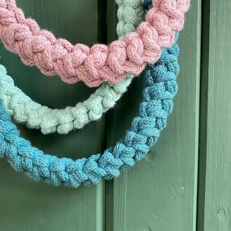 May include: Three braided rope wreaths in shades of pink, teal, and blue. The wreaths are made of thick, textured rope and are arranged in a layered composition against a green wooden background. The braided design is visible.