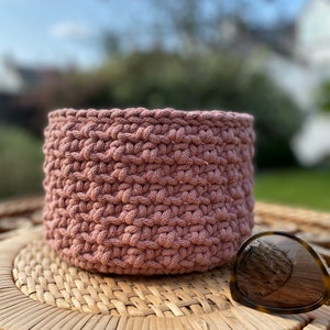 May include: A pink crocheted basket sits on a woven surface. The basket is made of thick cotton rope and has a round shape.