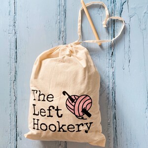 May include: A white canvas bag with a pink and black graphic of a yarn ball and crochet hook. The bag has the text "The Left Hookery" printed on it.