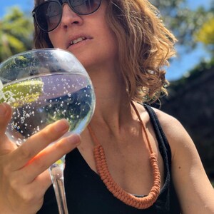 以下が含まれることがあります： サングラスをかけた女性と、錆色の織りネックレスのクローズアップ。 ネックレスは太い編み紐でできています。 彼女は泡の入った大きなグラスの水を手に持っています。
