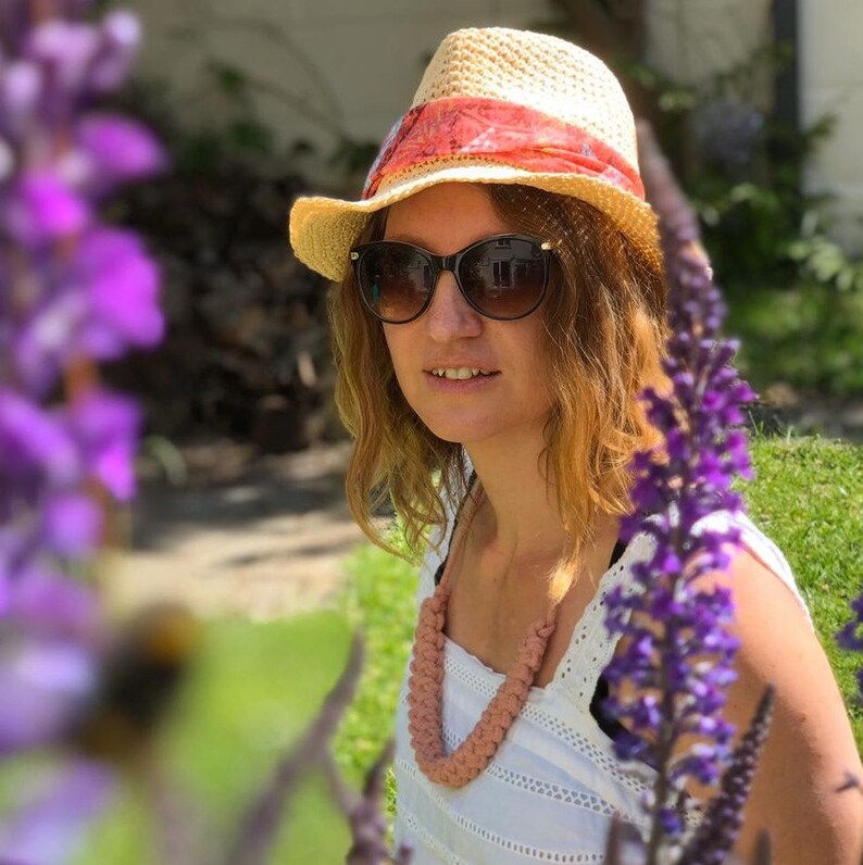 May include: A straw hat with a red and orange patterned band, worn with black sunglasses and a pink braided necklace. The hat has a wide brim and is slightly tilted. The necklace is a chunky, woven design.