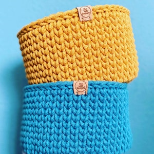 May include: Two crocheted baskets, one yellow and one blue, stacked on top of each other. Both baskets have a brown leather tag with the text "The Loft Hookery" on them.