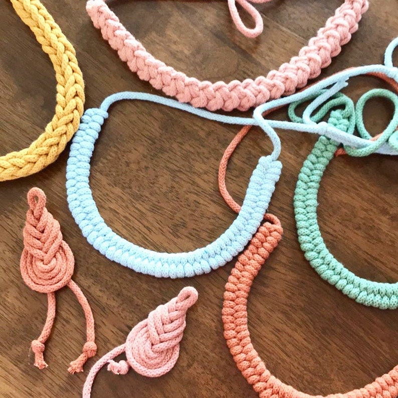 May include: A collection of handmade rope necklaces in various pastel colors, including yellow, pink, blue, and green. The necklaces are arranged on a wooden surface, showcasing their braided design and unique textures. Some necklaces have matching rope pendants.