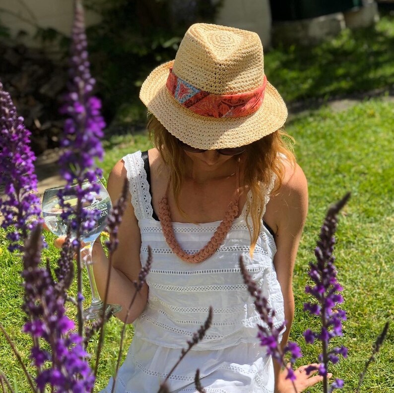 May include: A straw fedora hat with a patterned band, paired with a peach-colored woven necklace. The image is set outdoors with purple flowers in the foreground and a person wearing a white dress.