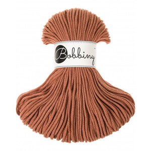 May include: A skein of terracotta-colored cotton cord, ready to be used for macrame or other crafts. The cord is bundled together and has a white label with the text "Bobbiny" in black.