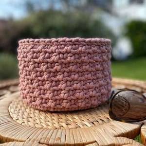 Puede incluir: Una cesta de crochet rosa polvorienta sobre una superficie de mimbre tejido. La cesta está hecha de cuerda de algodón gruesa y tiene una forma redonda.