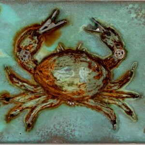May include: A ceramic tile with a rusty brown crab design on a teal blue background. The tile has silver metal accents around the edges.