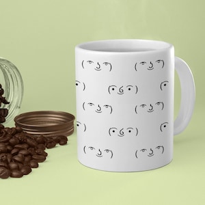 May include: A white ceramic mug with a pattern of black ( ͡° ͜ʖ ͡°) faces. The mug is filled with a steaming beverage and there are coffee beans scattered on a green surface.