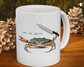 May include: White ceramic mug featuring a blue crab with a knife and the text "ask me again." The crab has red claws and is positioned as if wielding the knife. The mug has a white handle and is set against a blurred background.