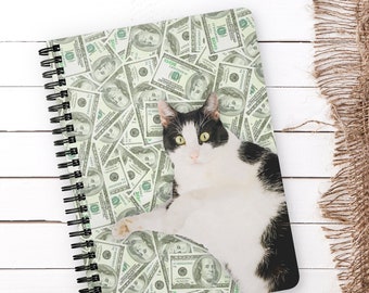 Puede incluir: Un gato blanco y negro con ojos verdes está tumbado sobre una libreta de espiral. La portada de la libreta está cubierta con un patrón de billetes de dólar estadounidense.