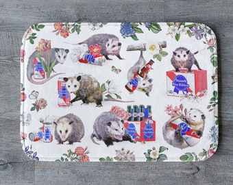 May include: White bath mat with a repeating pattern of opossums, flowers, and cans of PBR (Pabst Blue Ribbon) beer. The opossums are depicted in various poses, including holding cans of beer and sitting on top of cans of beer.