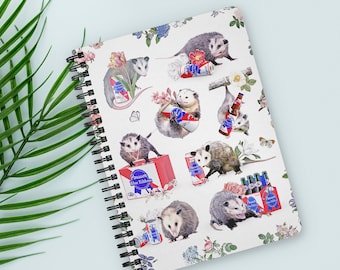 Puede incluir: Un cuaderno de espiral con un diseño caprichoso de zarigüeyas con latas y botellas de cerveza, rodeado de ilustraciones florales y mariposas. El cuaderno tiene un fondo blanco y una encuadernación en espiral negra.
