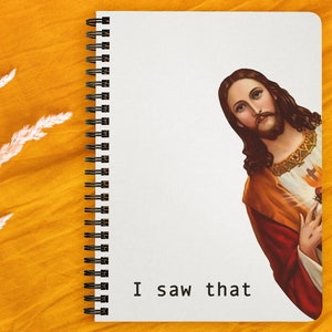 Puede incluir: Un cuaderno blanco de espiral con encuadernación negra. La portada presenta una imagen impresa de Jesucristo asomándose desde el lado derecho de la página. El texto "I saw that" está impreso en negro en la parte inferior de la página.