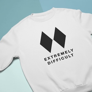 Puede incluir: Sudadera blanca con un gráfico de diamante negro y el texto "EXTREMELY DIFFICULT" impreso en la parte delantera.