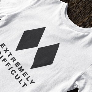 Puede incluir: Camiseta blanca con un gráfico de diamante negro y el texto "EXTREMELY DIFFICULT" impreso en la parte delantera.