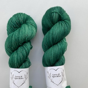 Könnte beinhalten: Zwei Stränge grüne Wolle mit weißen Etiketten mit einem Herz und dem Text "Love & Speckles". Das Garn ist ein leuchtender Grünton mit subtilen Farbvariationen, die ein strukturiertes Aussehen erzeugen. Das Garn ist fest gewickelt.