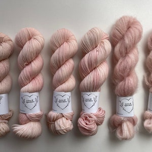 Puede incluir: Seis ovillos de hilo en varios tonos de rosa. El hilo está envuelto alrededor de tubos de cartón con una etiqueta blanca que dice "Love It Speckles".