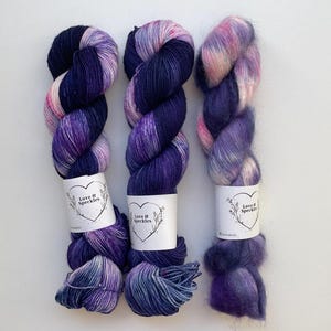 Op de afbeelding: Drie strengen handgeverfd garen in tinten paars, blauw en roze. Elke streng heeft een label met de tekst "Love & Speckles" in een hartvormig ontwerp. Het garen is gerangschikt op een wit oppervlak.