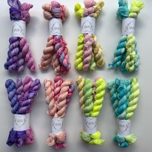 May include: Eight skeins of colourful yarn in shades of purple, pink, yellow, and teal. Each skein is wrapped with a white label reading "Love & Sparkles". The yarn is arranged on a white surface.
