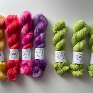 May include: Four skeins of yarn in various colors, including yellow, pink, purple, and green. The yarn is wrapped around cardboard tubes with labels that say "Love & Sparkles".